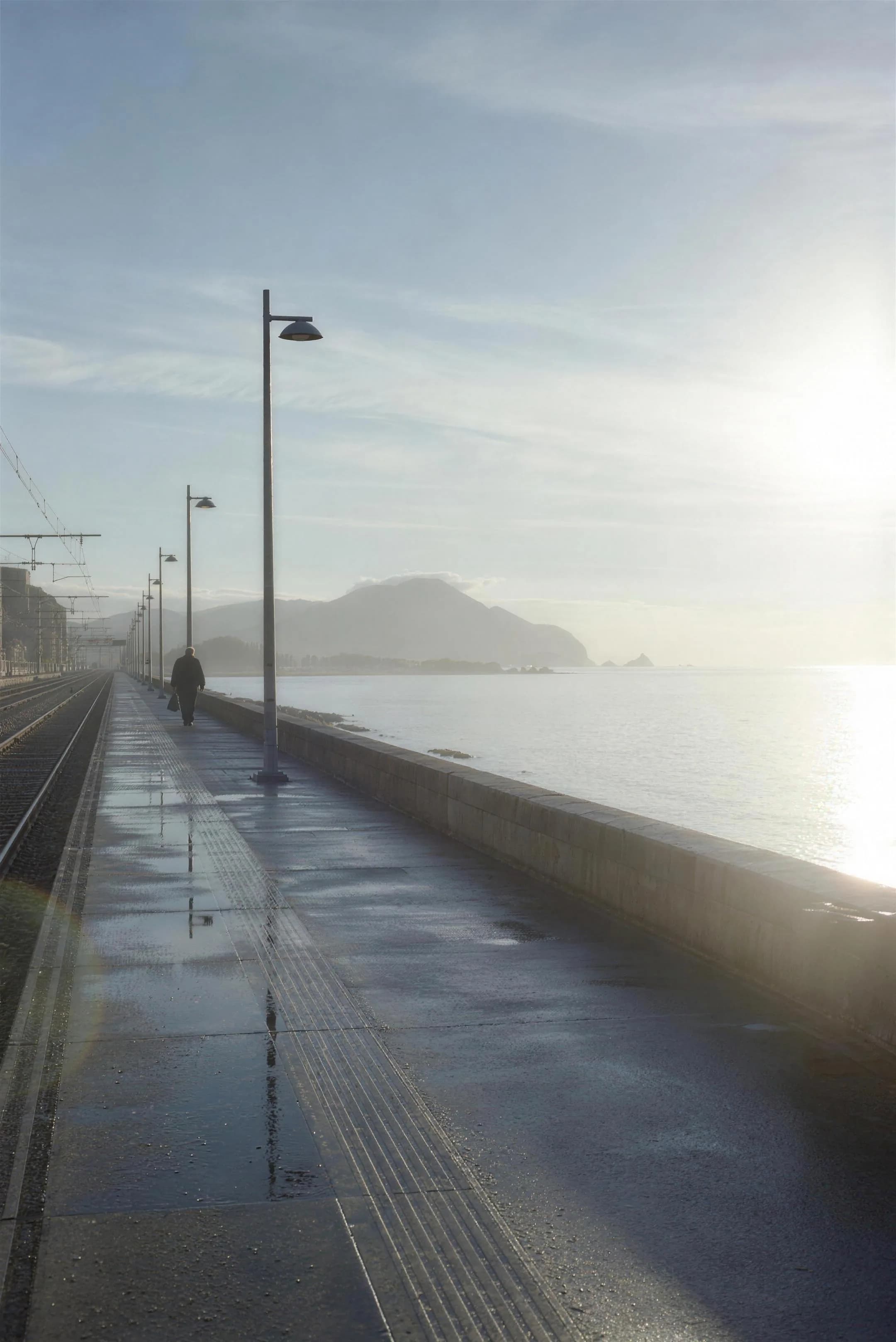 An empty seafront platform in winter morning light, a lone figure with a bag far down the platform, low sun flaring gently off the sea