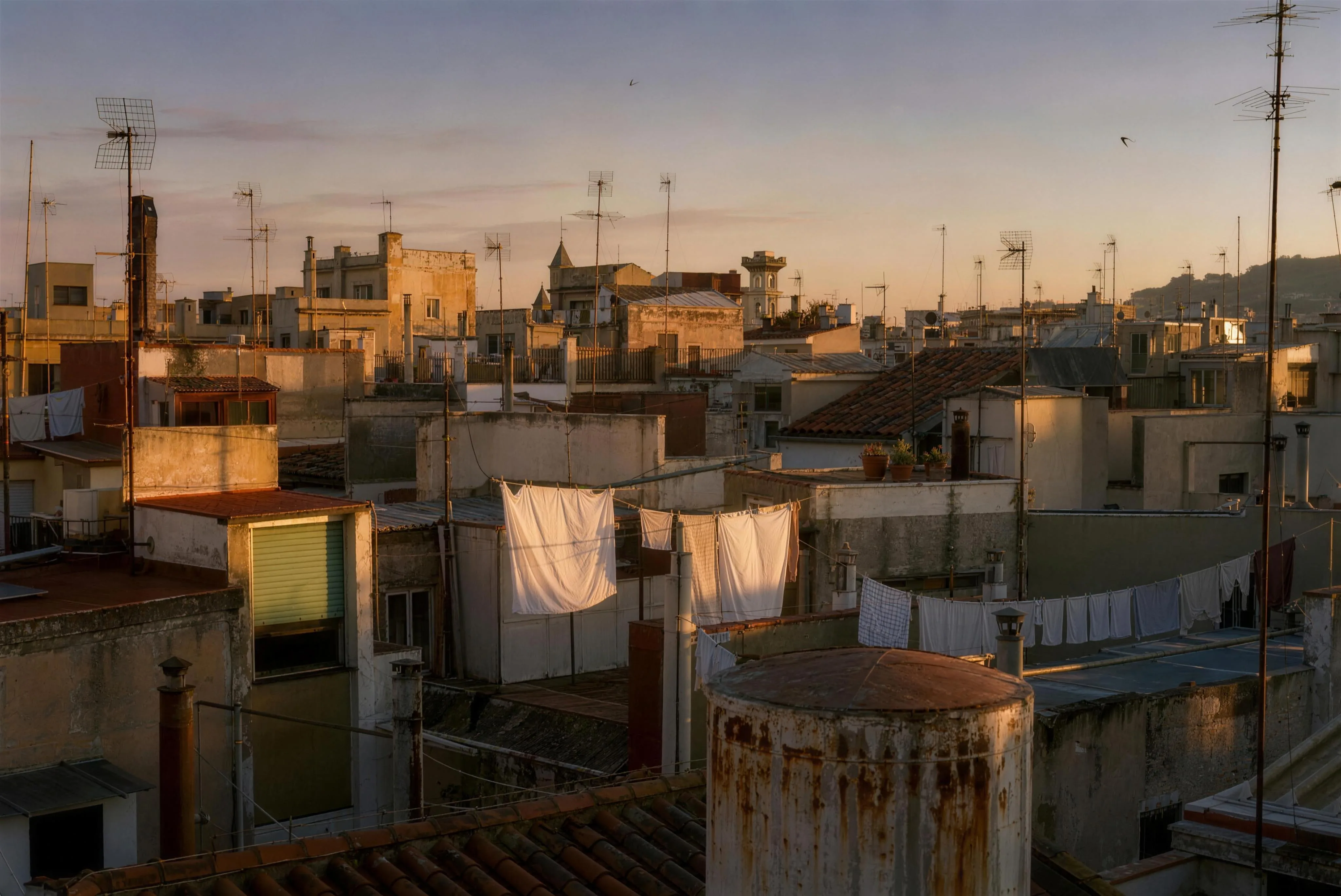 A sea of Barcelona rooftops in the last hour of evening light — terracotta tile, backlit laundry, water tanks and television aerials against the warm sky